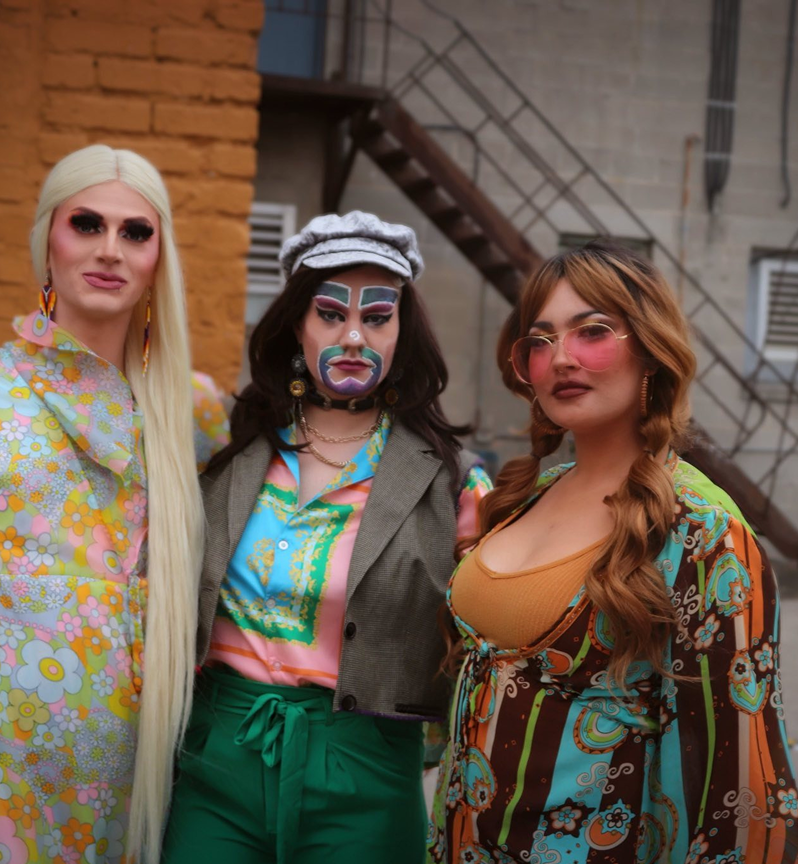 Three drag performers with elaborate makeup and colorful retro outfits pose outdoors.
