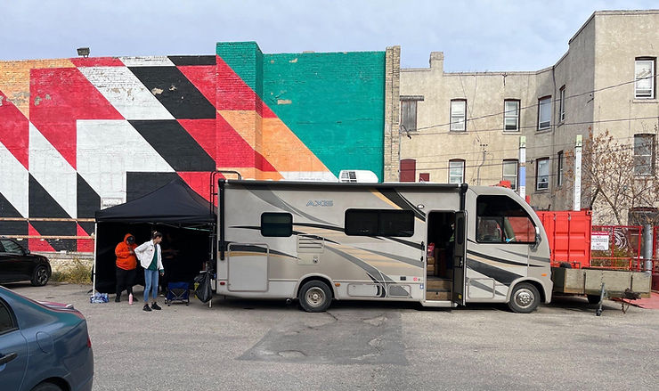 A photo of the MOPS RV parked in a lot. Behind the RV is an apartment building and a large wall painted with a mural that looks like a star blanket. Behind the RV is a black tent, and in front of the tent are two MOPS staff members.